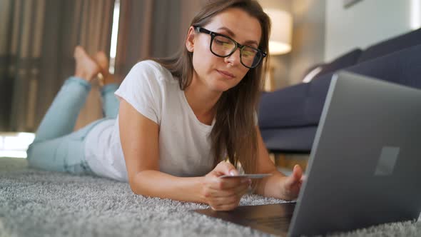 Woman with Glasses Is Lying on the Floor and Makes an Online Purchase Using a Credit Card and Laptop