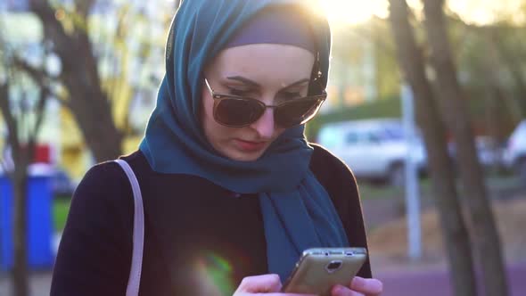 Portrait of a Girl in Hijab Uses Phone at Sunset in Park