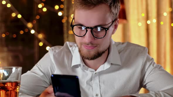 Handsome Bearded Man Using a Smartphone and Smiling While Drinking Beer in Pub