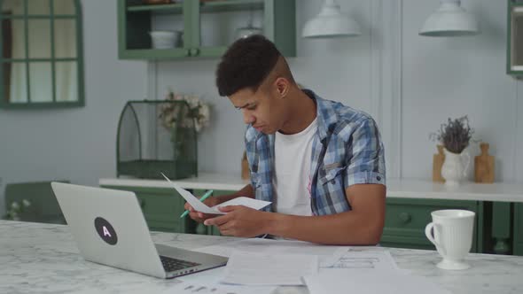 20s African American Man Working with Paper Documents Using Laptop Sitting in Kitchen