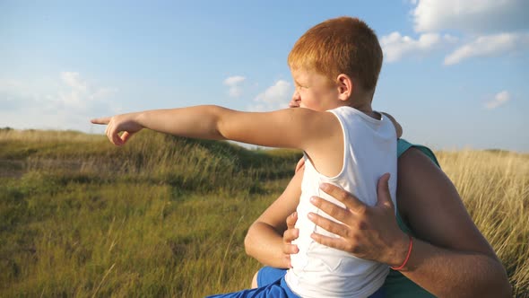 Little Son Sitting at Green Grass on the Lawn with His Dad and Pointing at Something to Him