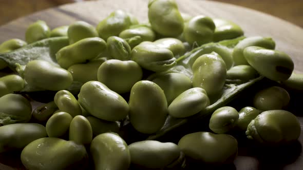 Pile of rotating sweating, backlit, green fava beans piled on top of a couple of favas in their pods