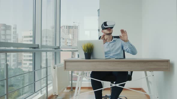 A Young Man Sitting at a Desk in the Office Uses Augmented Reality Glasses To Work on Business