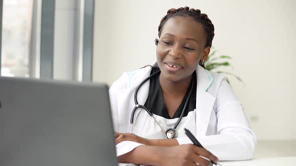 Young African American Woman Doctor Having Chat or Consultation on Laptop and Makes Some Notes