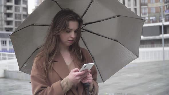 Unhappy Concentrated Woman Typing on Smartphone As Standing Outdoors with Umbrella. Portrait of Sad