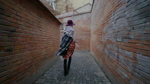 Female tourist joyful walking and slightly jumping passing a bricked wall street in unknown city.