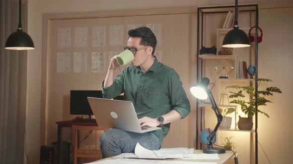 Designer Sits On His Desk Holds Laptop On The Knees Drinks Coffee While Working On The Project