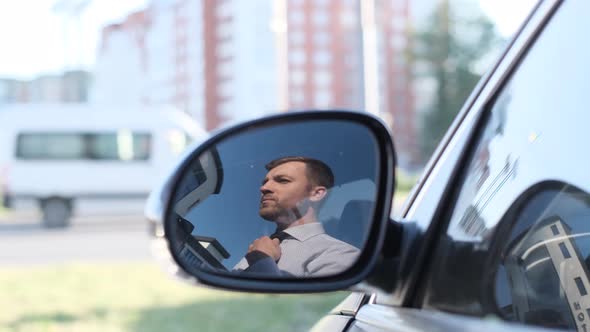 Young Businessman Man Puts on a Tie on the Street Looking Out the Window of His Car