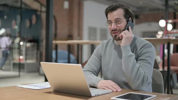 Young Man Talking on Smartphone While Using Laptop in Office