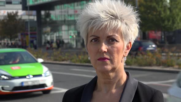 A Middle-aged Woman Looks Seriously at the Camera in an Urban Area - Closeup - a Busy Road