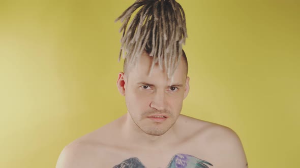Naked serious young man with tattoo on chest chewing gum on yellow background.