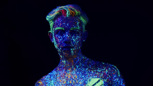 Young man painted with glowing colors looks into camera do gesture of silence, front view. Man paint