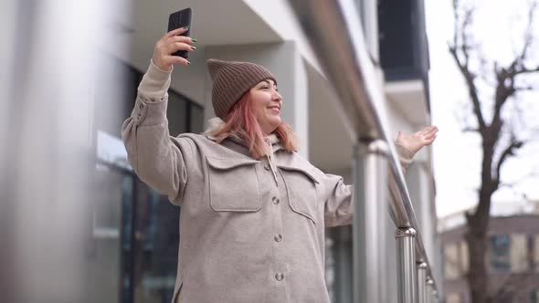 Shooting From Below of Cheerful Overweight Female Vlogger in Warm Outerwear and Hat Recording Video