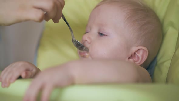 Woman Feeds Funny Son with Baby Fruit Puree in Highchair