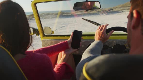 Happy caucasian couple sitting in beach buggy by the sea using smartphone