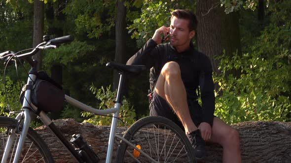 A Young Handsome Cyclist Sits on a Log Next To His Bike in a Forest and Talks on a Smartphone