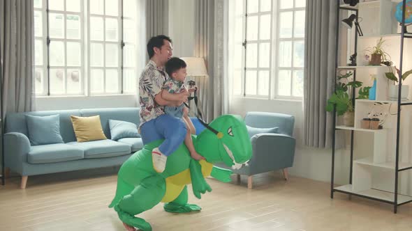 Asian Father Carrying His Son While Ride Dinosaur Toy In Modern House Living Room, Happy Family