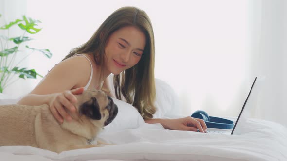 Woman typing and working on laptop with dog Pug breed lying on bed