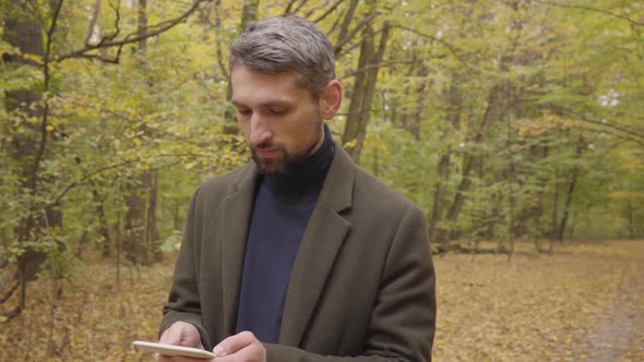 Handsome Caucasian Man Standing in the Autumn Forest with Tablet and Looking Around