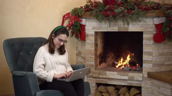 A Young Pregnant Woman with Glasses Sits in an Armchair By the Fireplace with Horns on Her Head and