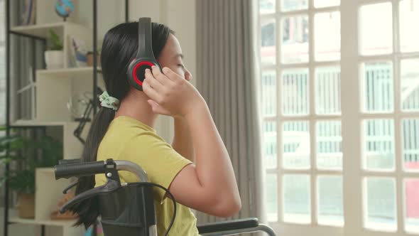 Asian Kid Girl Sitting In A Wheelchair Listening To Music And Looking Out Of A Door