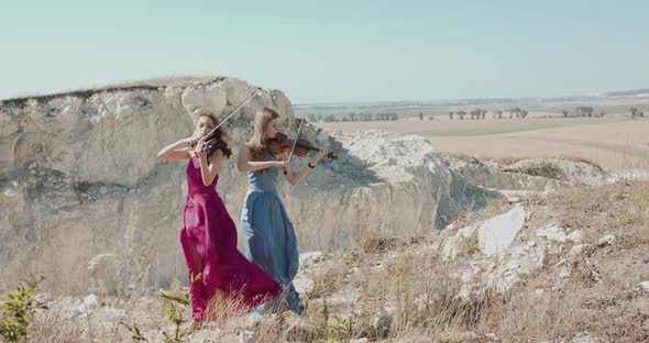 Female Duet in Elegant Blowing Dresses Plays the Violin Among Picturesque Rocks