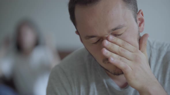 Close Up Portrait of Crying Man on the Foreground and His Shouting Angry Wife Sitting on the Sofa