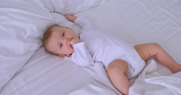 Pretty Baby with Big Blue Eyes is Lying on a White Bed and Sucking His Fingers