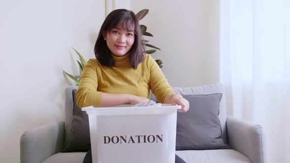 Portrait of Asian young woman looking at camera holding box with clothes for donation. Concept campa