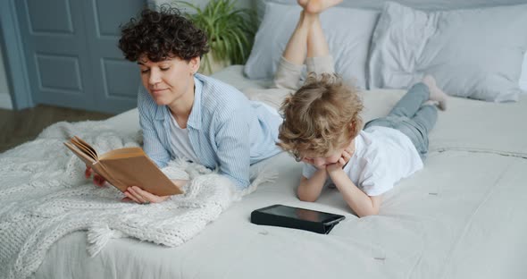 Little Boy Watching Cartoons on Tablet While Mother Reading Book in Bed at Home