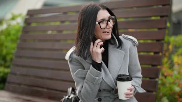 a Brunette in a Gray Coat Glasses and a Paper Cup in Hand is Sitting on a Highbacked Wooden Bench