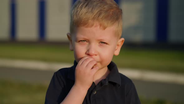 Little Boy Touches Nose and Smiles Standing on City Street