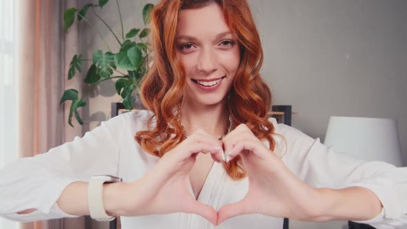 Happy Redhead Caucasian Woman Millennial Smiles and Shows Heart Made of Fingers