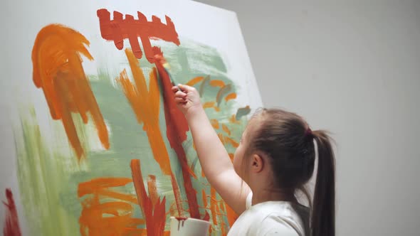 Kid Girl with Down Syndrome Draws with a Brush on a Large Canvas in a White Room Girl with Special