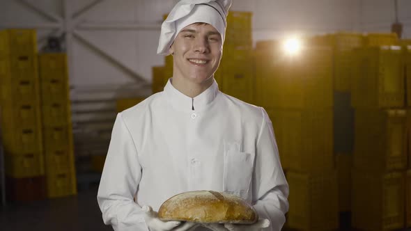 Happy Baker in Uniform Holds a Fresh Loaf of a Bread in a Bakery with Backlit