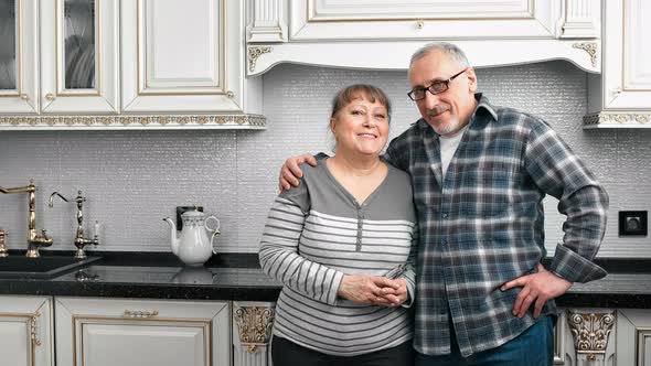 Smiling Beautiful Elderly Woman and Man Hugging Smiling Posing in Kitchen Looking at Camera