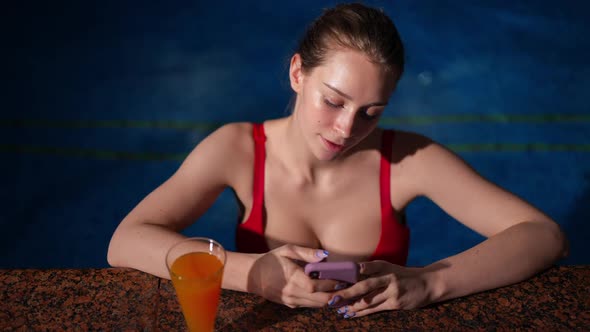 Front View Relaxed Young Woman Messaging on Smartphone at Poolside with Orange Cocktail