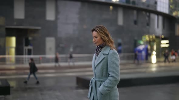 a Blonde in a Light Coat Has Hands in Pockets and is Walking Thoughtfully in the Twilight Against