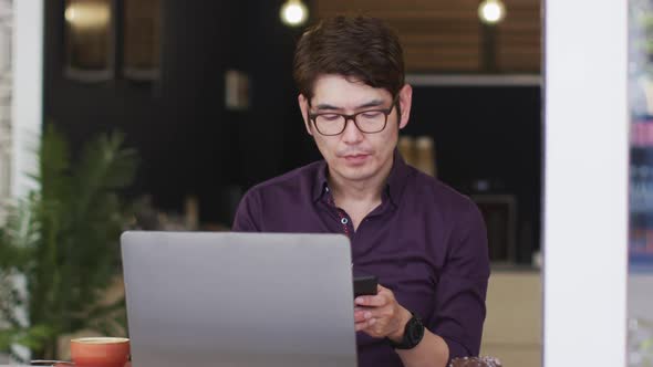 Asian man with laptop talking on smartphone while sitting at a cafe