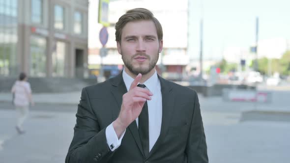 Outdoor Portrait of Young Businessman with Finger on Lips Please Be Quiet