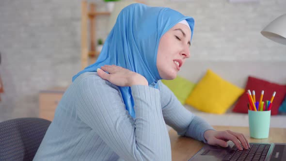 Young Muslim Woman in a Traditional Headscarf Slouches While Sitting at a Laptop and Feels Pain in