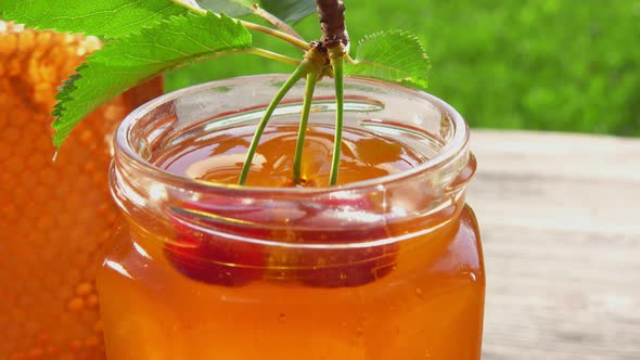 Two Large Cherries on the Twig with Leaves are Dipped Into Fresh Honey in a Jar