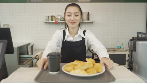 Portrait of smiling Asian woman barista people in apron serving a cup of coffee and croissants