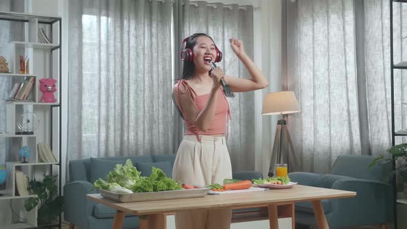 Happy Asian Woman Listening To Music With Headphones And Singing While Slicing Cucumber At Home