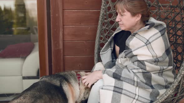 A woman plays and petting a shepherd dog while sitting in a wicker chair in a country house.