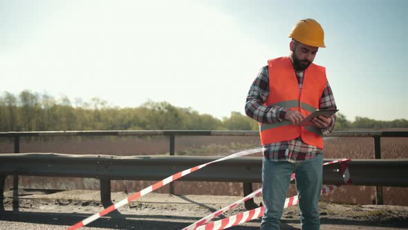 Young Bearded Man Inspector in an Orange Vest and Protective Helmet with Tablet