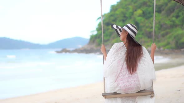 Back of Young sexy Asian woman with big striped hat swings on seafront swing relaxing on tropical is