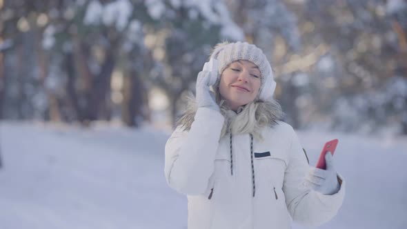 Portrait of Cute Girl in Warm Winter Clothes Listening to Music in Big White Headphones Using a