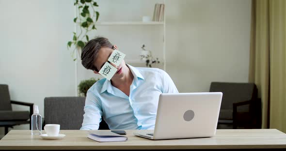 Tired Comic Male Manager Pretends Working Sleeping with Stickers on Face Sits at Desk with Laptop