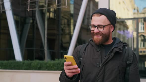 Joyful Man in Glasses Looking at Phone Screen and Smiling While Walking at Street. Handsome Bearded